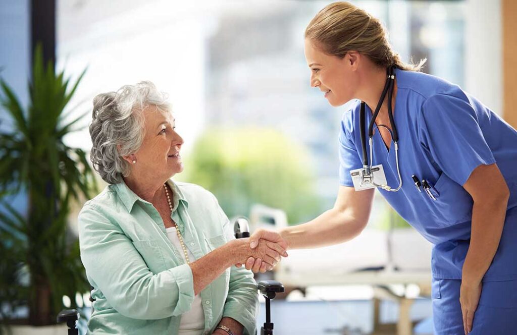 An older adult chats with friendly staff and residents in a bright assisted living common room.
